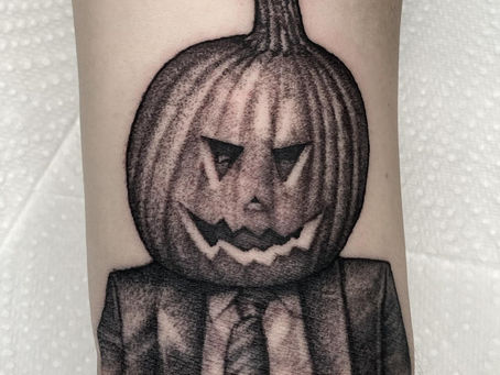 Tattoo of a pumpkin head wearing a suit and tie on skin, featuring jagged eyes and smile. Background is textured, likely a paper towel.