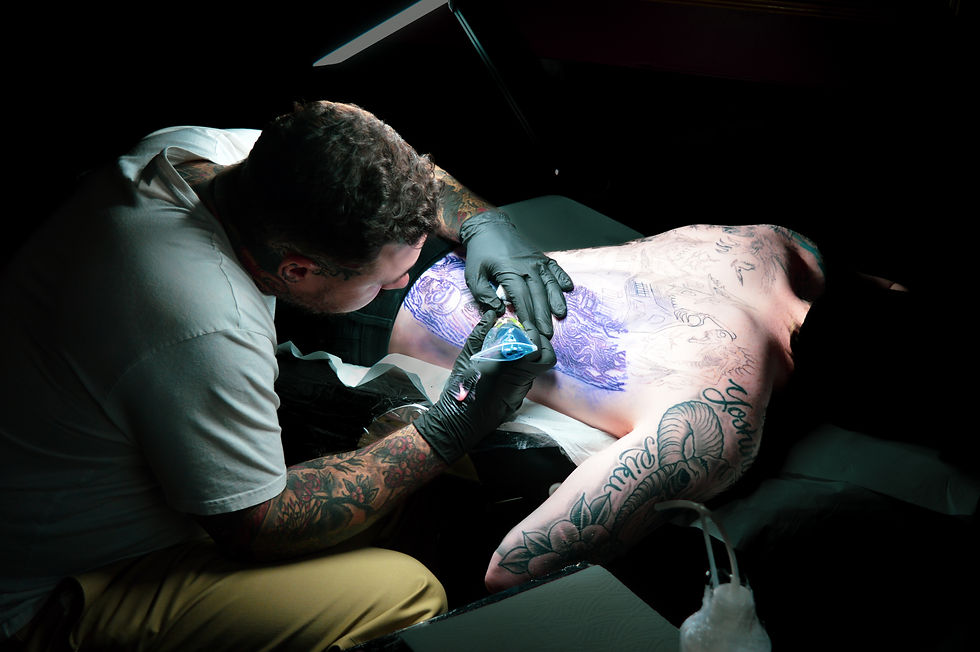 Tattoo artist in white shirt and gloves inking intricate blue design on a man's back. Dimly lit studio with visible tattoos on skin.