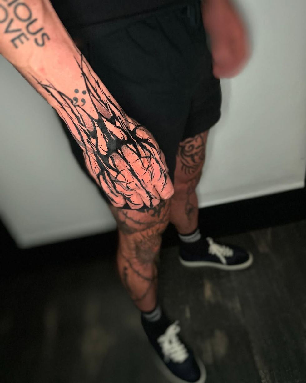 Person with intricate black tattoos on hand and legs, wearing black shorts and sneakers. The word "LOVE" visible on the arm.