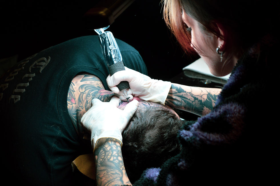Tattoo artist with tattooed arms works on a man's head in a dimly lit setting. The man wears a black shirt with text.