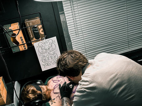 Tattoo artist in white shirt creates back tattoo on seated client. Ring light illuminates scene. Sketches and blinds in background.