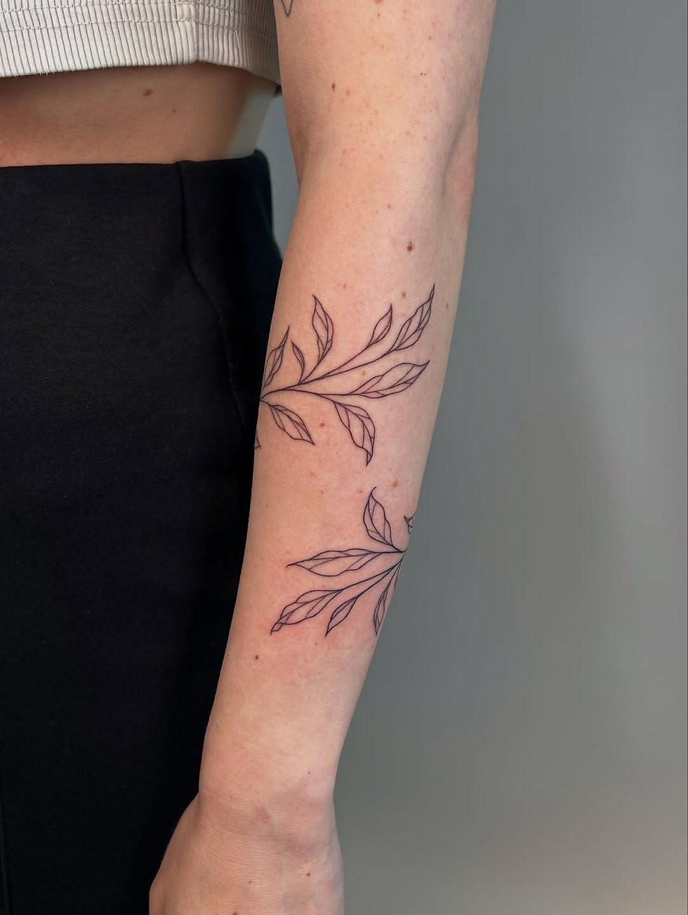 Arm with black line-art leaf tattoos shown, wearing a beige top and black pants, set against a plain gray background.