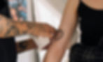 Tattoo artist with tattooed arms applies a heart design on a woman's arm. The tattoo reads "Make Yourself Hard to Kill". Indoors, casual setting.