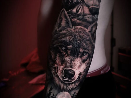 Tattoo of a wolf, moon, and forest on a person's arm in dim red lighting. The design is detailed, conveying a mysterious and intense mood.