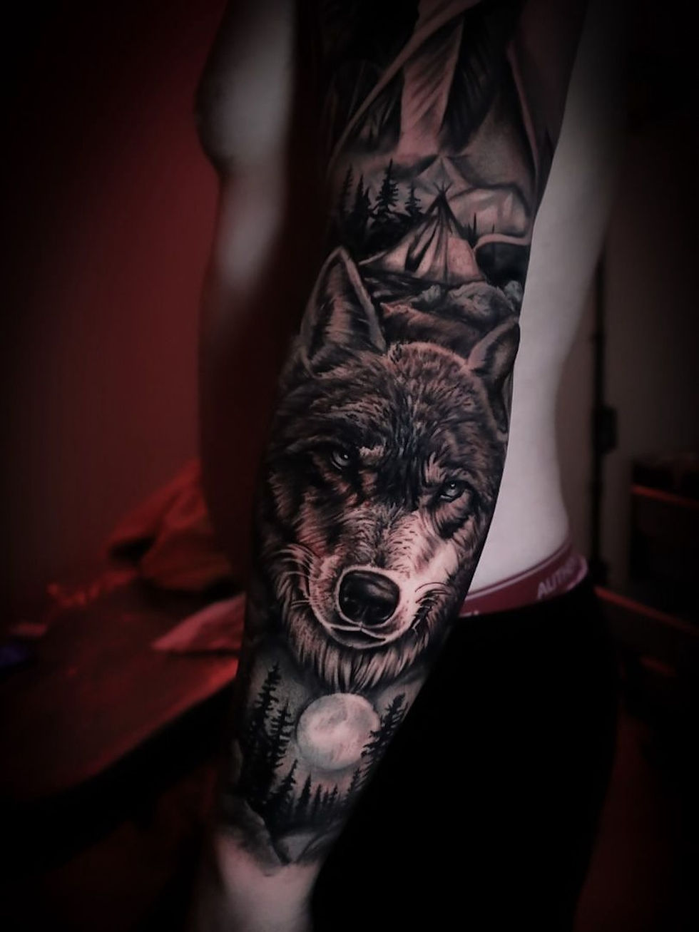 Tattoo of a wolf, moon, and forest on a person's arm in dim red lighting. The design is detailed, conveying a mysterious and intense mood.