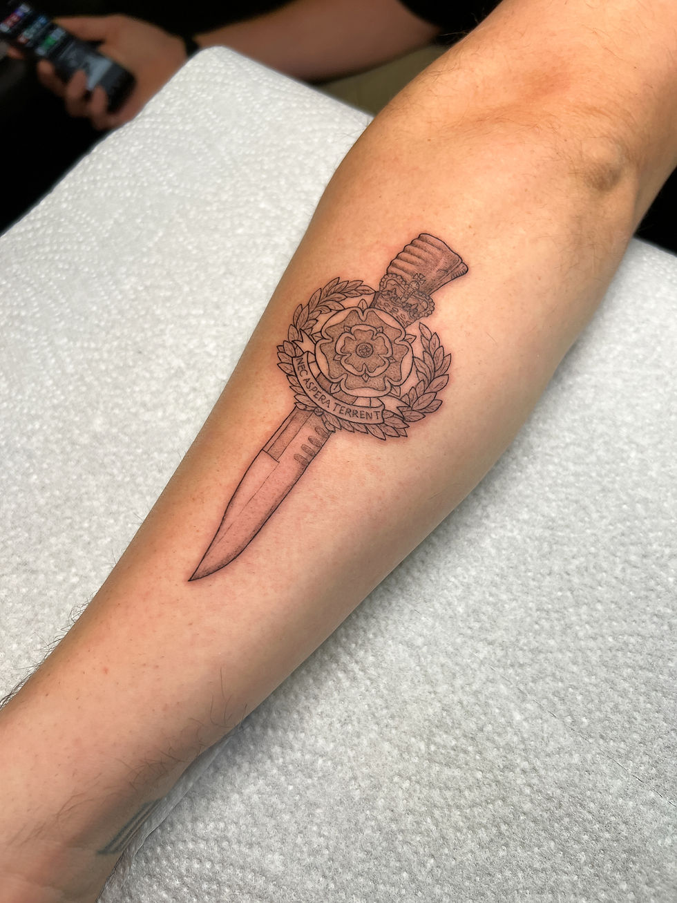 A person in a white tank top shows a tattoo on their arm in a mirrored room. Green plant leaves are visible in the foreground.