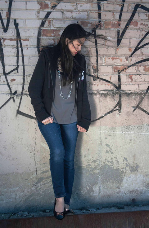 A portrait of a  teen in a black sweater stands in front of a brick, graffiti filled wall as the wind blows her black hair around her face.