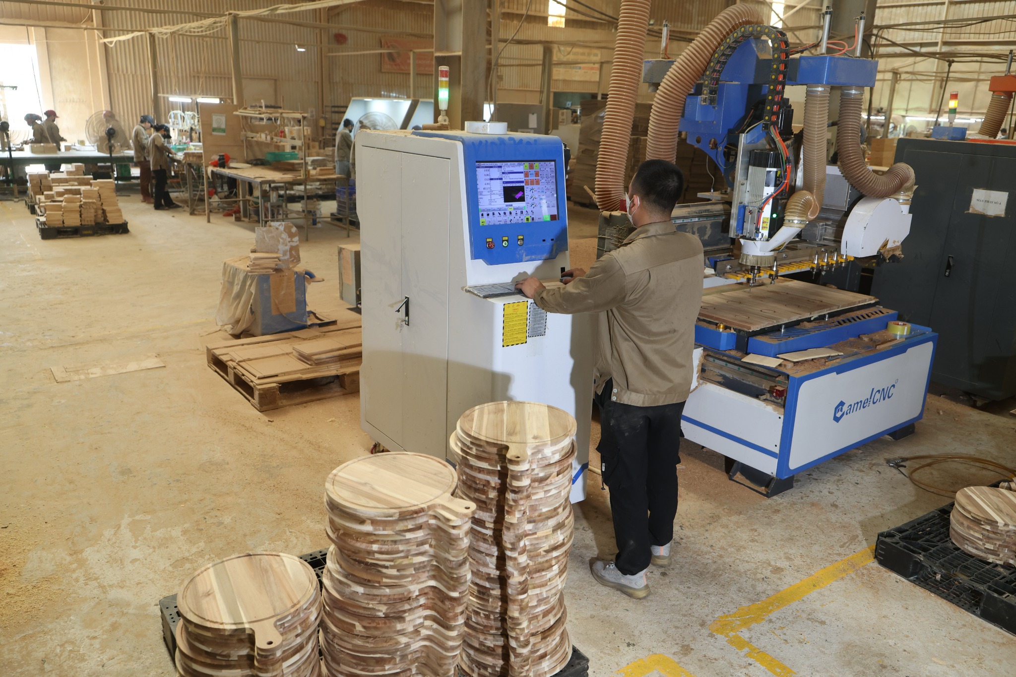 Factory team member using CNC controls with woodworking staff and stacked serving boards in the Vietnam workshop