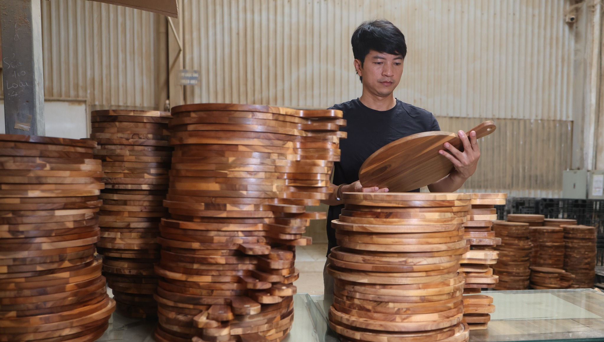Factory worker inspecting a wooden paddle board beside stacks of completed boards