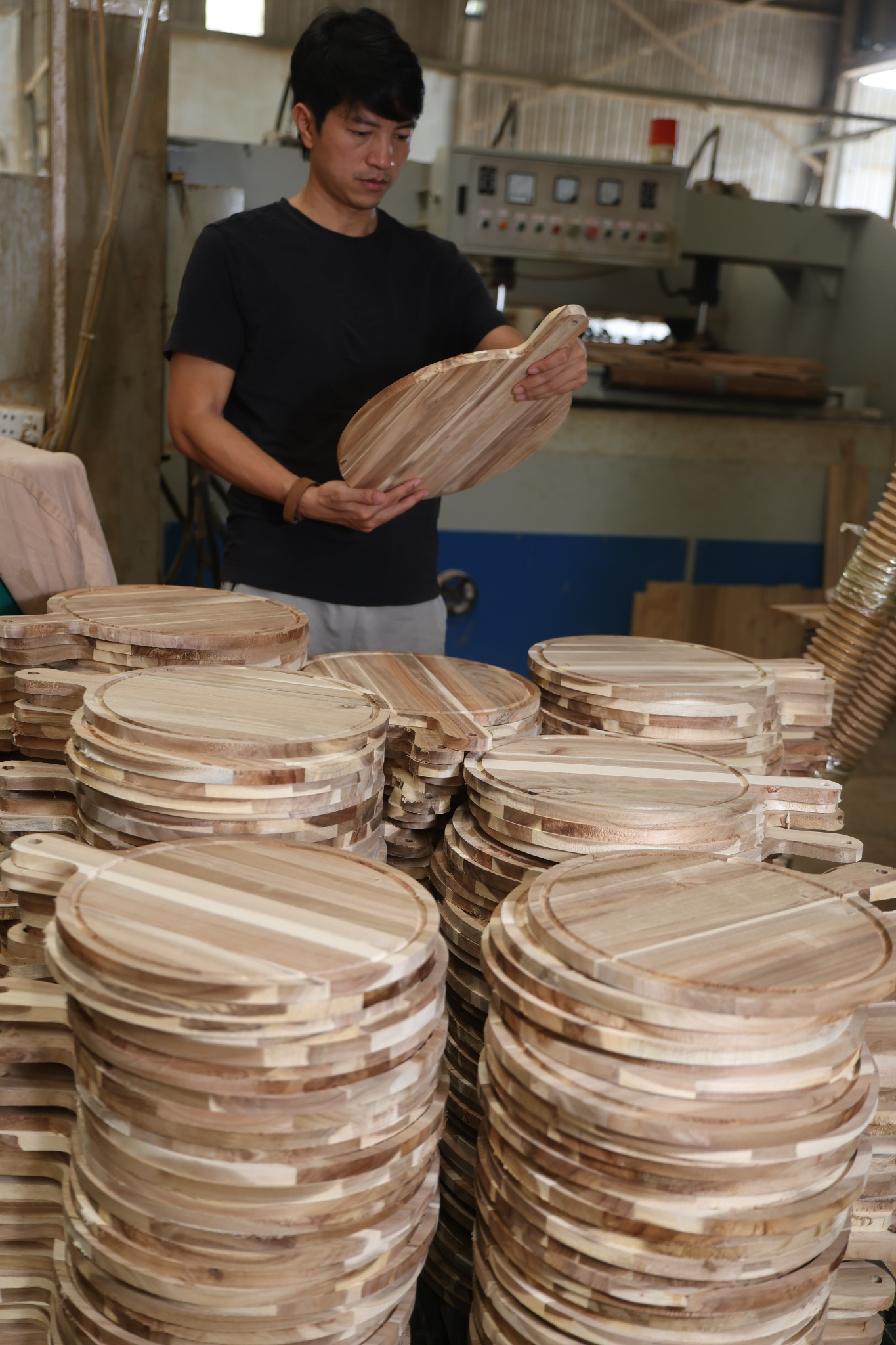 Factory worker inspecting a round wooden paddle cutting board beside stacked boards in Vietnam