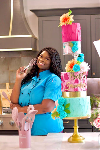 A professional food photography shot of a Black woman baker in a bright turquoise chef's coat standing in a modern kitchen. She is holding a silver whisk and has long, natural curly black hair. Behind her is a tall, multi-tiered wedding cake decorated in hot pink and mint green with turquoise blue roses and coral-colored flowers. The cake has three tiers with different designs - the top tier is hot pink with a coral flower topper, the middle tier features mint green frosting with turquoise roses, and the bottom tier combines both colors with gold accents. The kitchen background shows gray cabinets and a stainless steel range hood. In the foreground, there is a pink utensil holder with kitchen tools. The lighting is warm and professional, creating soft shadows and highlighting the vibrant colors of the cake and chef's uniform. The image has a professional food photography quality with sharp focus and rich color saturation.