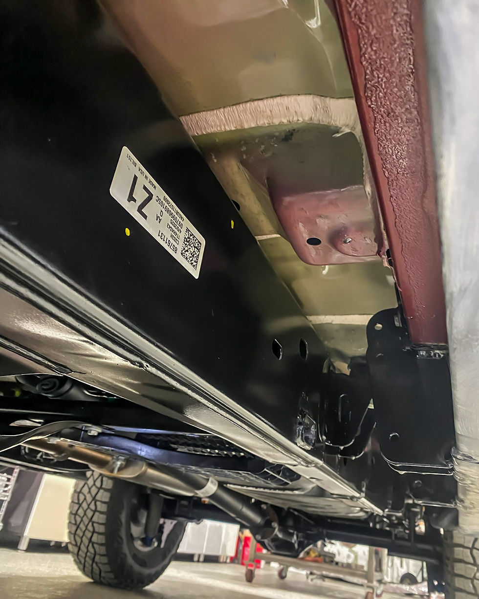 Underside of a vehicle on a lift, showing black undercoating, metal frame parts, tire, and a white label marked 21.