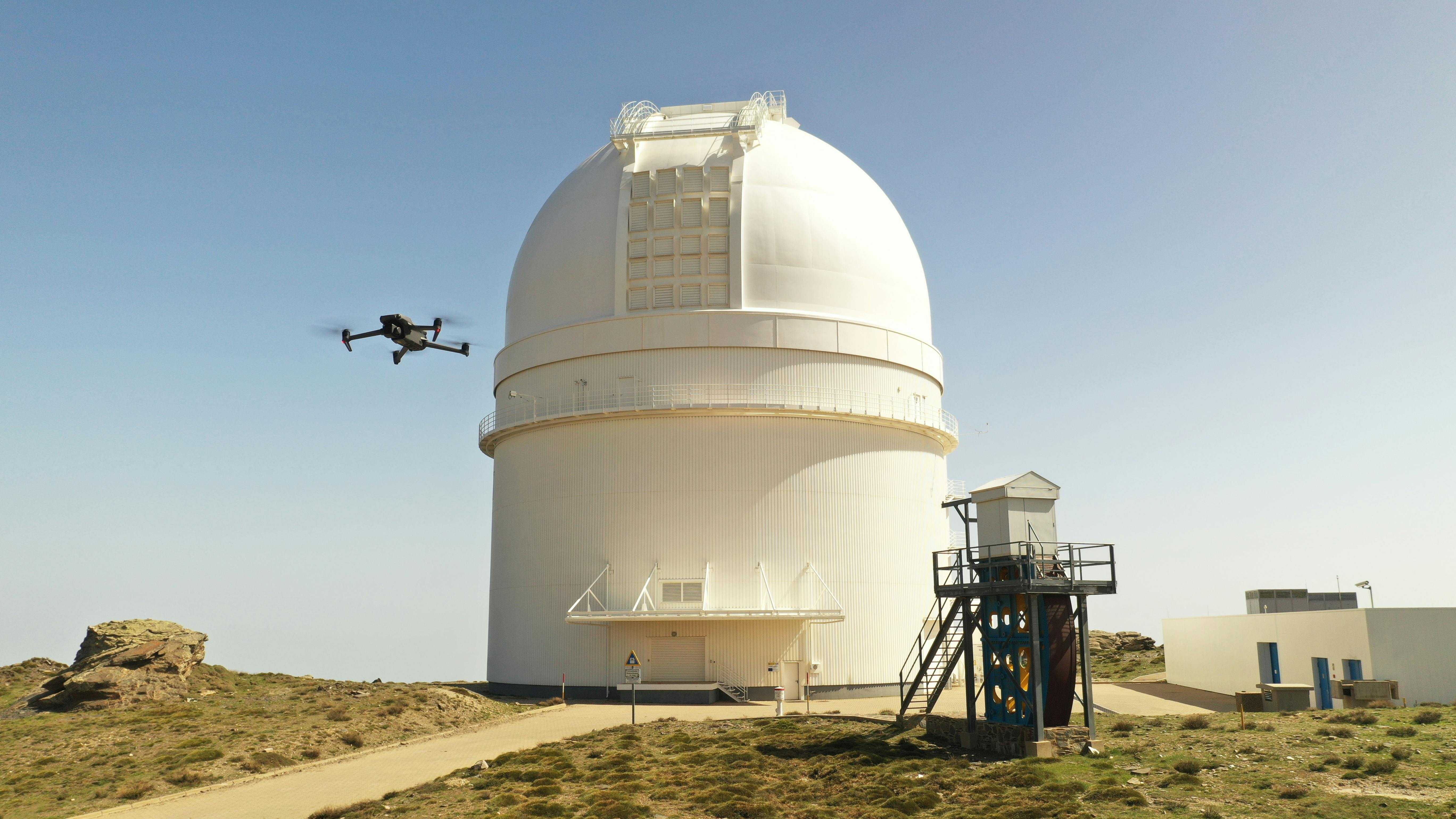 Drone flying near large white observatory dome on a sunny day.