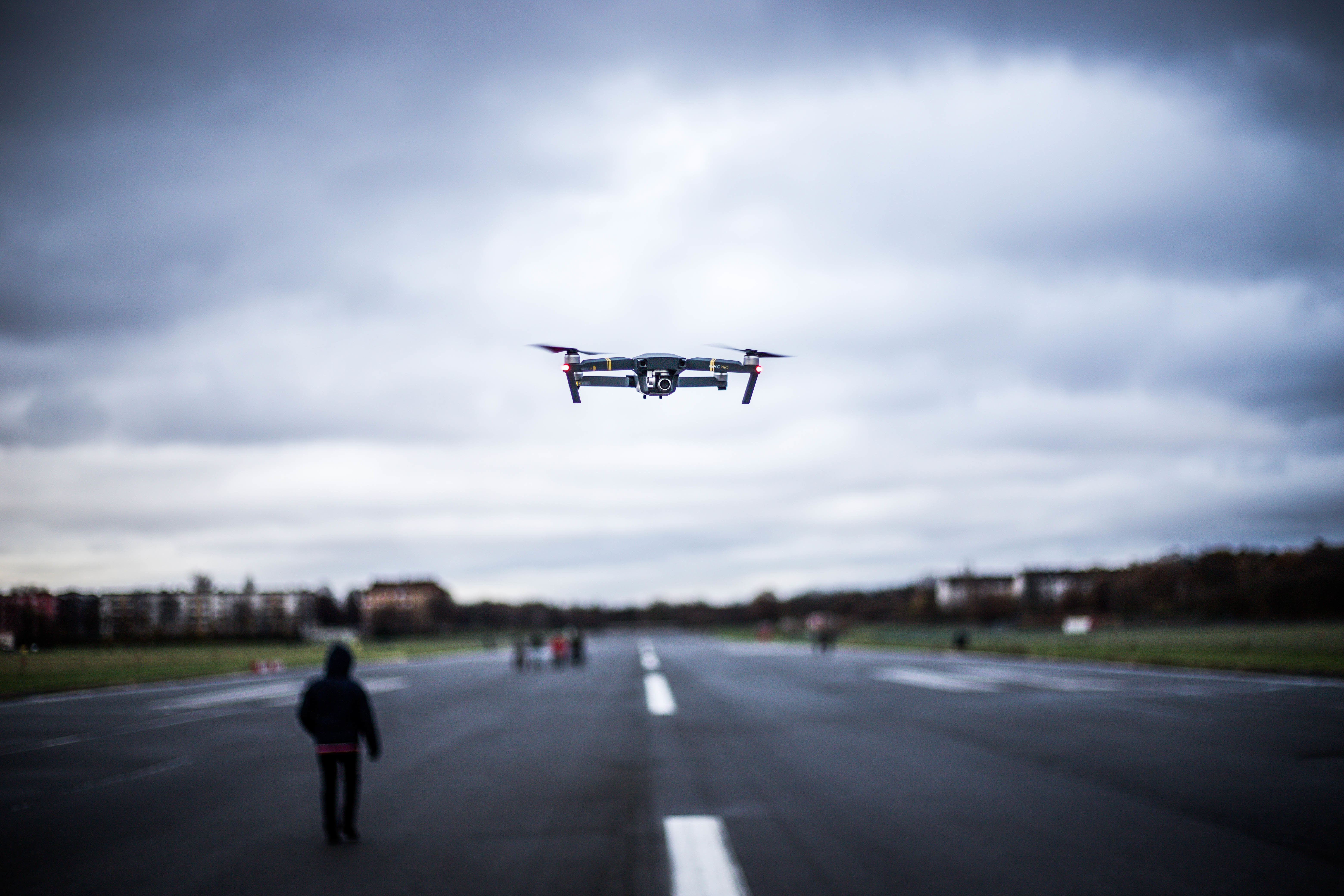 Drone flying overhead, person walking on runway, cloudy sky background.