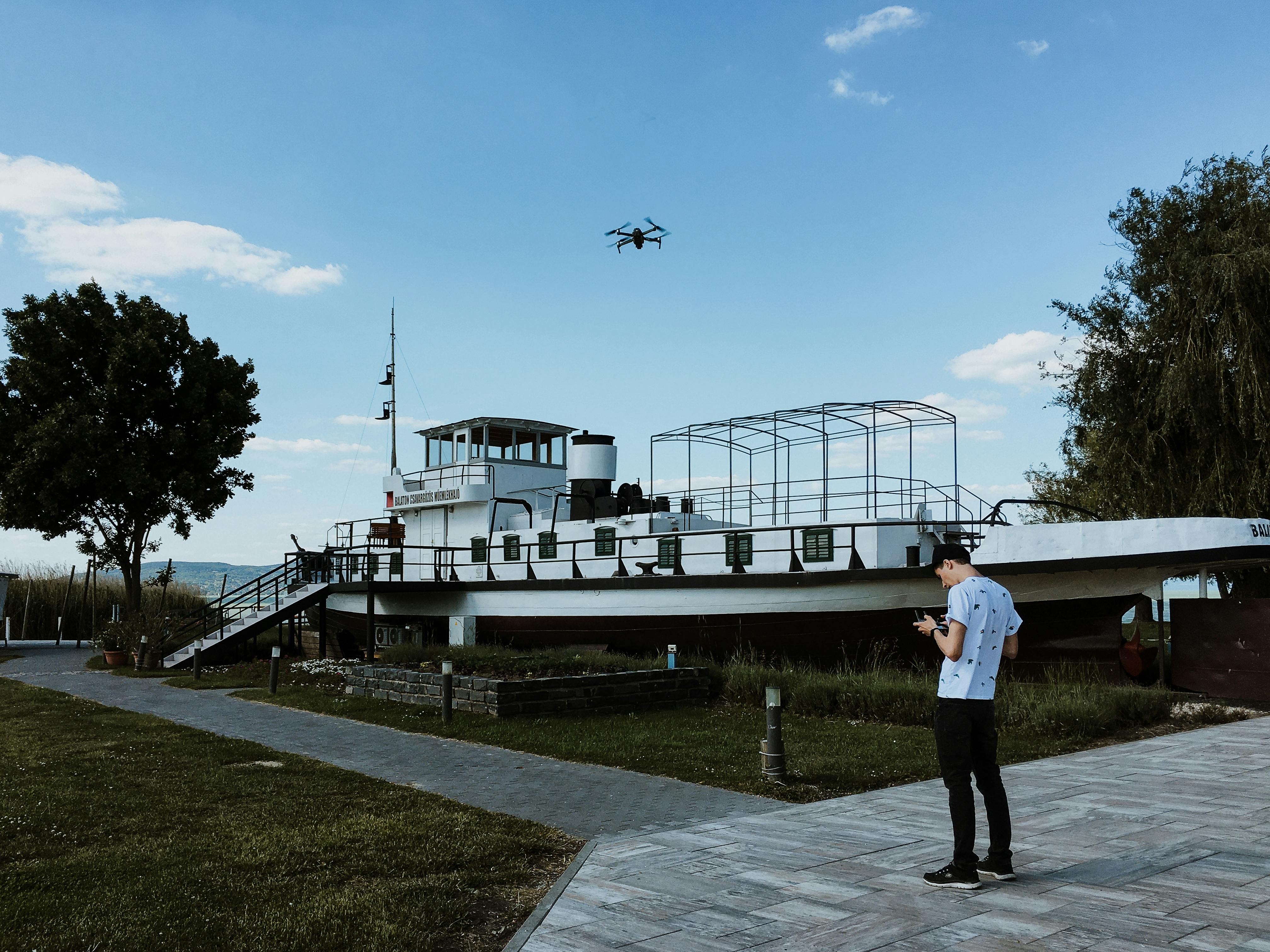 Man operating drone near boat, blue sky, and waterfront building. Pilot