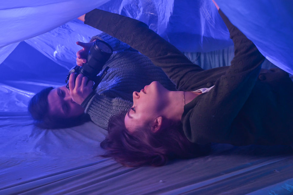 Eine Schauspielerin und eine Kamerafrau liegen unter einer Plastikplane und drehen eine mystische Szene.