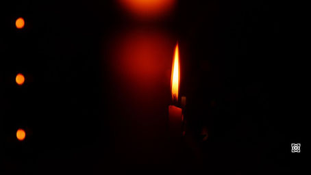 Single lit candle flame glows orange in a dark room, with soft red and orange bokeh lights; moody, quiet scene.
