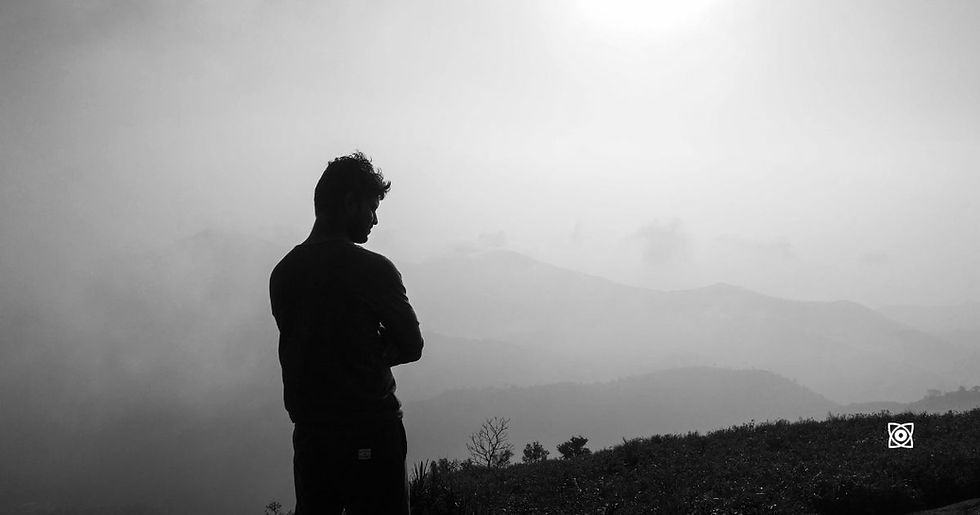 Silhouette of a man standing with arms crossed on a hill, overlooking misty layered mountains in black and white.