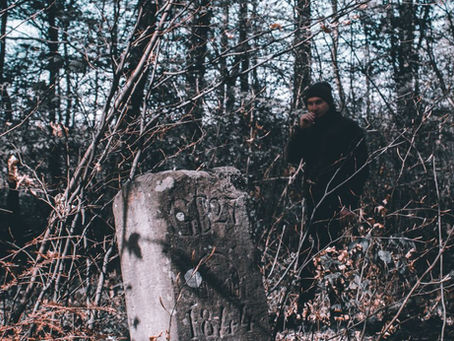 A weathered gravestone in a forest, with a person standing behind it and smoking.