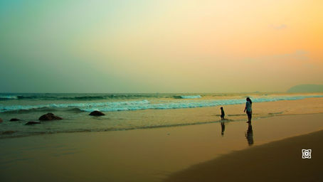 A woman and child stand on a quiet beach at sunset, with gentle waves, rocks, and a warm orange sky.