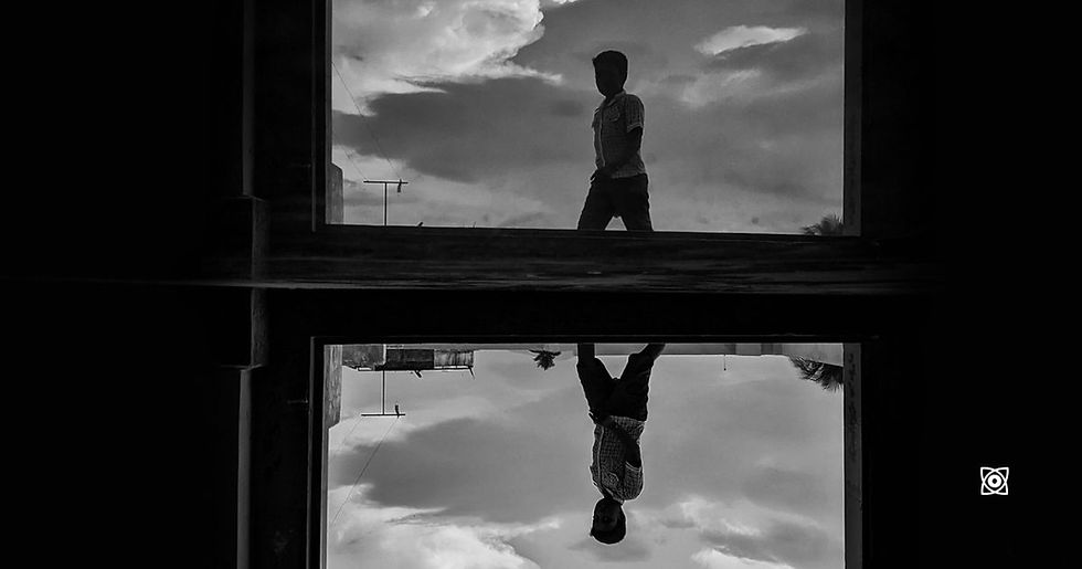 Black-and-white photo of a boy walking past a window, with his upside-down reflection below and cloudy sky behind a dark frame.
