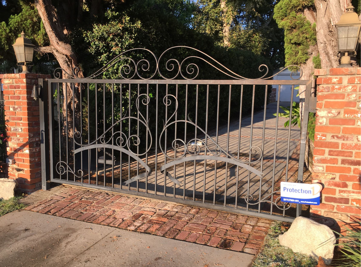 Black iron driveway gate with scroll top between red brick pillars over a brick paver driveway in Moorpark, CA