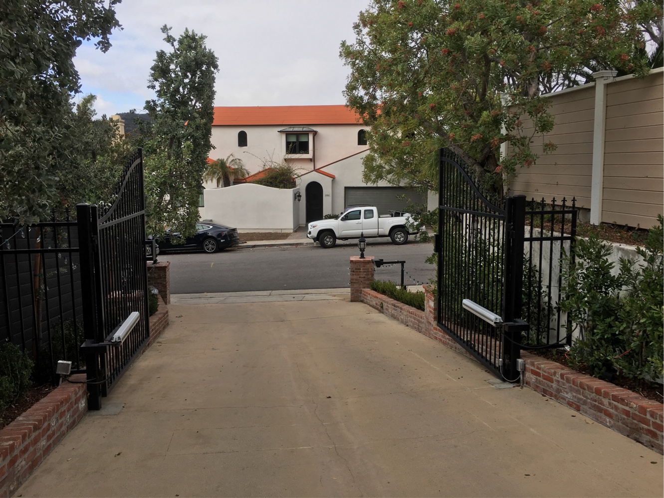 Black iron double swing driveway gate open at a residential entrance with brick planters in Northridge, CA