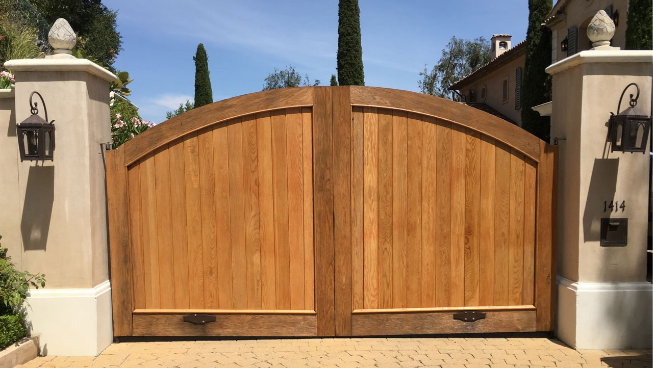 Arched natural wood double driveway gate between stucco pillars with lanterns in Camarillo, CA