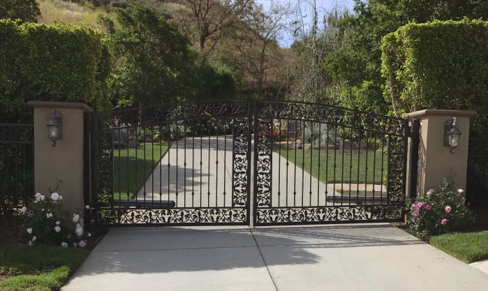 Black wrought iron double driveway gate with ornate scroll base between stucco pillars in Tarzana, CA
