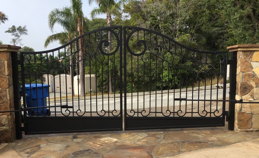 Arched black wrought iron double driveway gate with scroll top between stone pillars in Arleta, CA