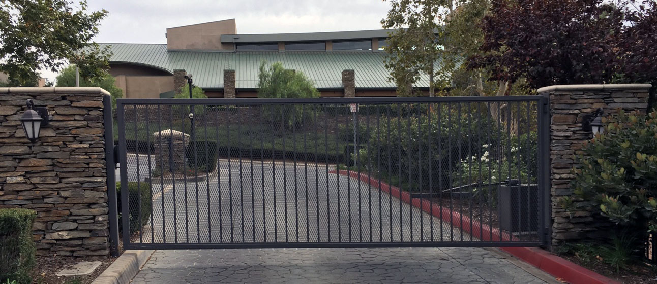 Black iron slide gate between stacked-stone pillars with lanterns at a community entrance in Porter Ranch, CA