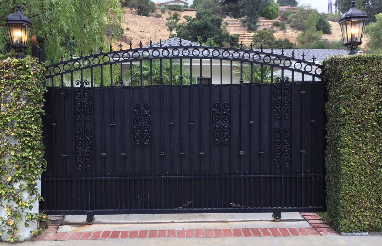 Arched black solid-panel driveway gate with scroll top between hedges on a brick driveway in Granada Hills, CA