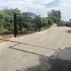 Arched black iron driveway gate across a long dirt ranch road lined with orchards in Somis, CA