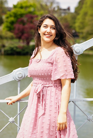 Merced is standing on a bridge on a sunny day. She is wearing a pink dress and looking at the viewer.
