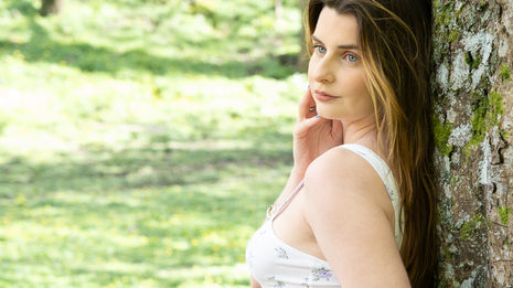 Woman with long hair leans against a tree, gazing thoughtfully outdoors.