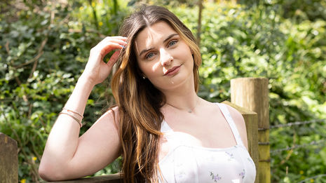 Young woman with long brown hair smiles by wooden fence outdoors.
