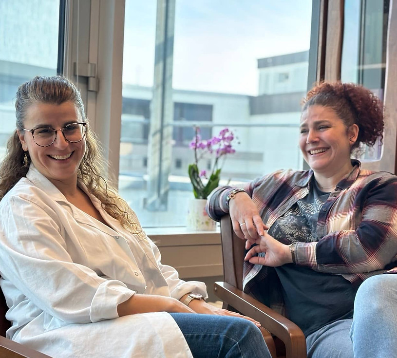 E. Agelonia und Maria sitzen gemeinsam in einem hellen Praxisraum und lachen.
