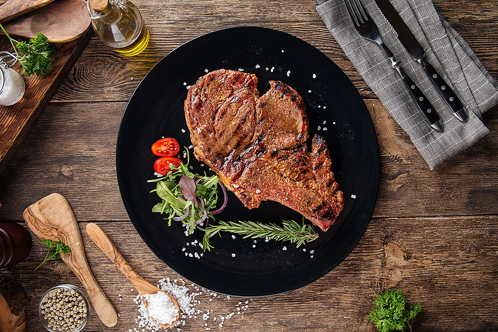 Entrecôte cuite dans une assiette, photographie vue de haut. L'assiette est noire, posée sur une table en bois. Sur la photo, il y a également des cuillère en bois, de l'huile, du sel, des herbes aromatiques, un torchon, des couverts.