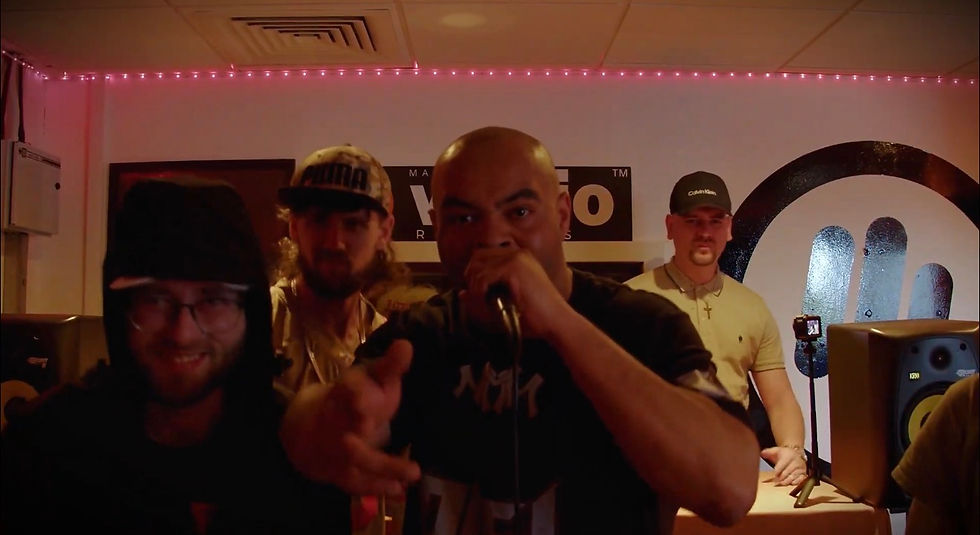 Four men in a studio, one rapping into a microphone as others stand behind him under pink string lights.