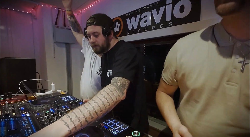 Three men at a DJ booth under a Wavio Records sign, one mixing on a controller, with pink lights in the studio.