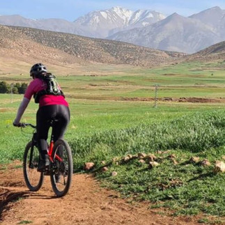 A person rides a bike on a dirt path through the Kik Plateau, with the Atlas Mountains visible in the background.