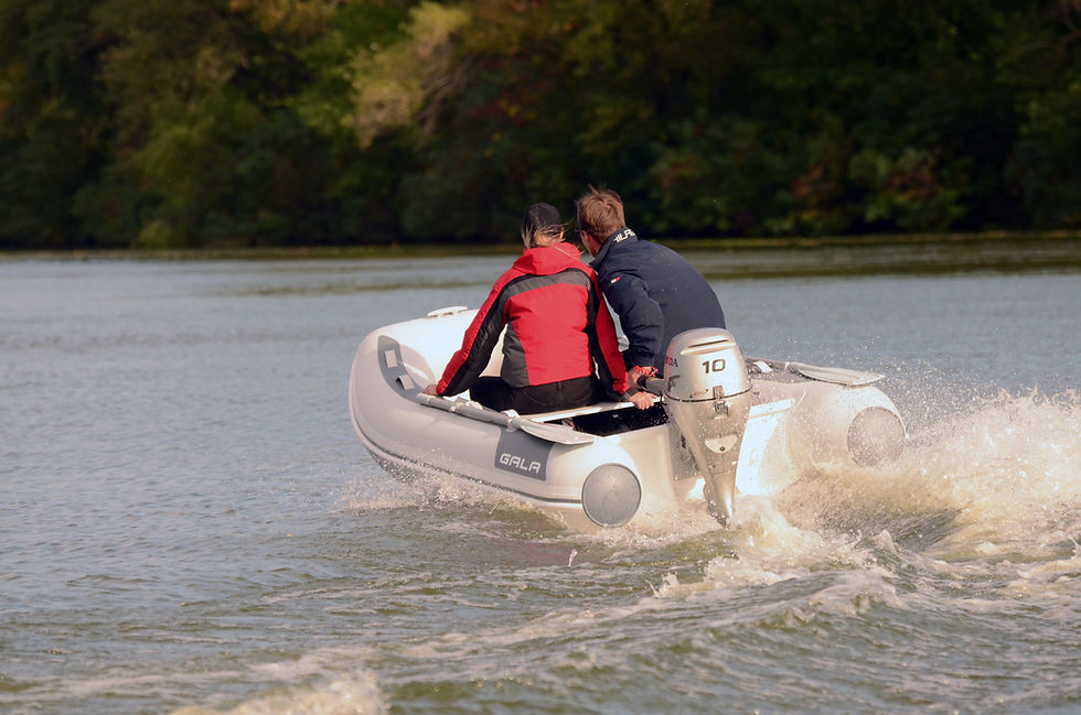 Two people ride a small white inflatable motorboat labeled GALA across a lake, splashing water, with green trees behind. A great rib tender for sale from GALA.