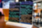 A pint of amber beer and a chalkboard menu listing various beers and ciders with colorful text on a wooden table in a cozy bar setting.