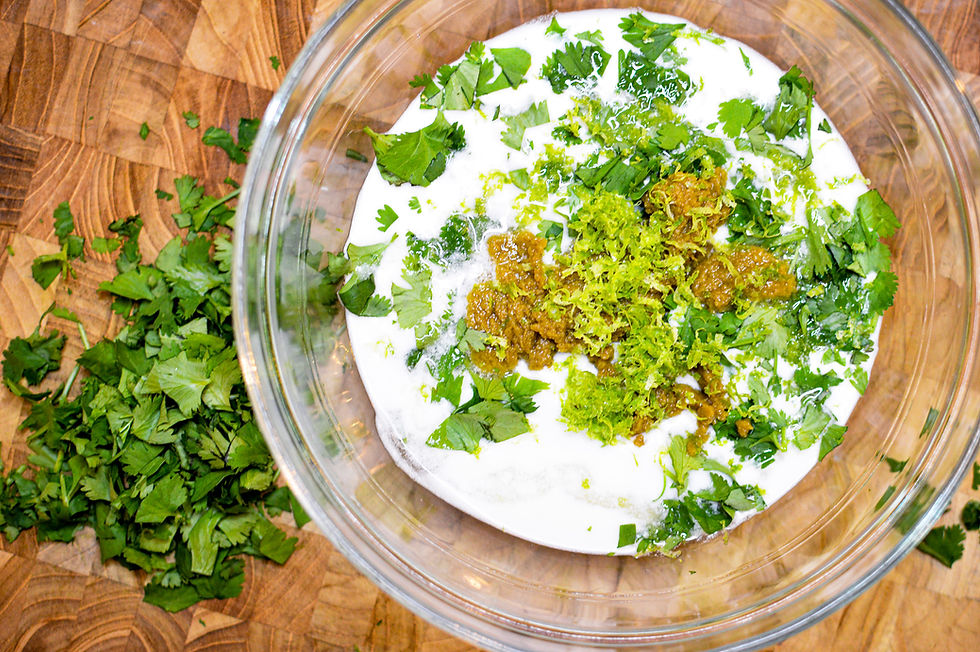A glass bowl with yogurt, chopped cilantro, zest, and spices on a wooden surface; fresh cilantro on the side.