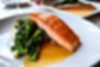 Seared salmon with broccolini and rice on a white plate, drizzled with sauce. Blurred background with a second plate. Warm, appetizing.