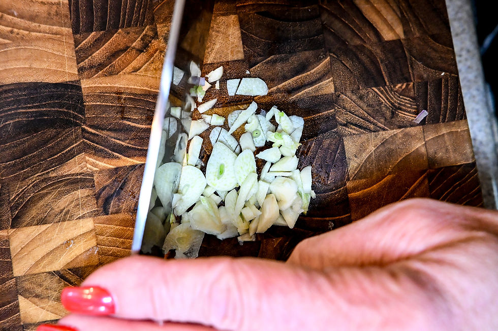 Close-up of chopped garlic on a patterned wooden cutting board, with a knife and a hand with red nail polish nearby.