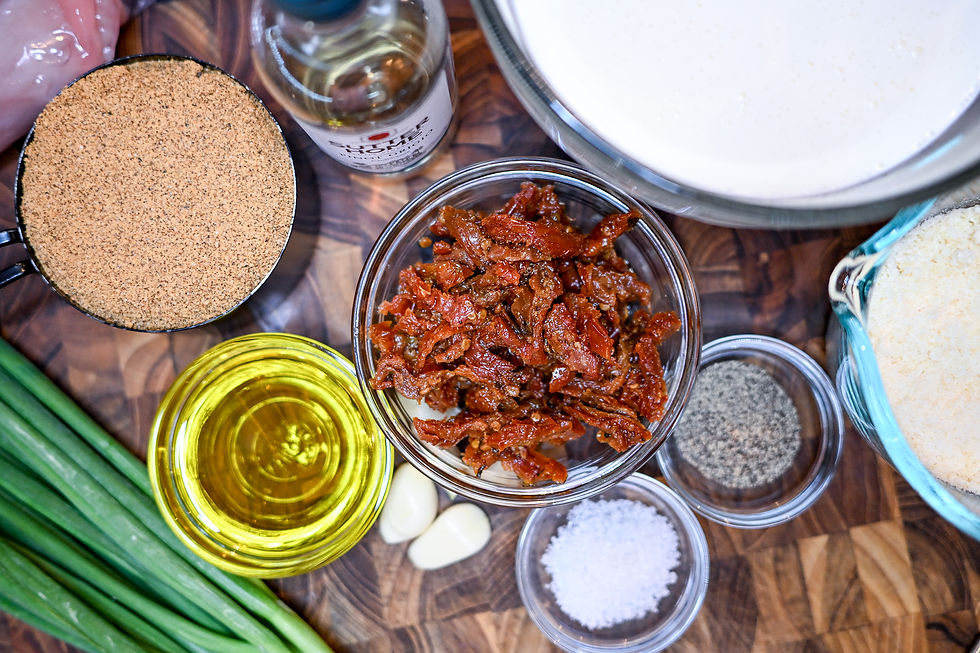 Ingredients on a wooden board: sun-dried tomatoes, breadcrumbs, oil, salt, pepper, garlic, cream, green onions, chicken, and a small bottle.
