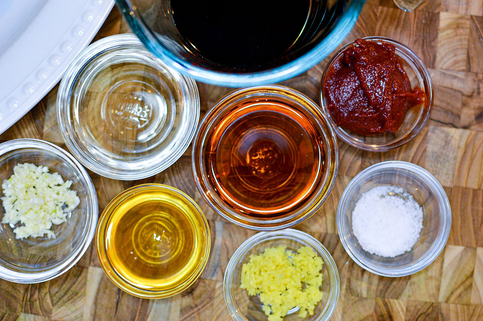 Various spices and sauces are arranged in small glass bowls on a wooden surface, including garlic, ginger, soy sauce, and tomato paste.