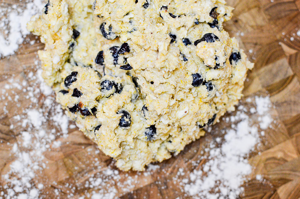 Close-up of uncooked dough with black olives on a floured wooden surface. Dough appears lightly textured and sprinkled with flour.