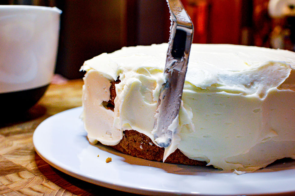 A knife spreads creamy white frosting on a cake on a white plate. A cup sits nearby on a wooden table, creating a cozy kitchen scene.